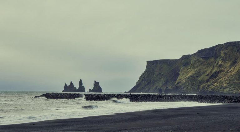 Waves on the black sand beach of Reynisfjara, with sea stacks offshore and a mossy cliff under an overcast sky